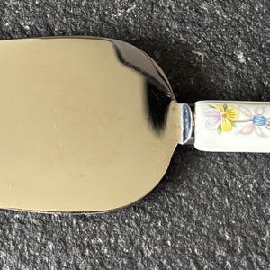 Può includere: Una paletta per torte in acciaio inossidabile con un manico a fantasia floreale. La lama è di colore argento lucido e il manico presenta una base bianca con fiori blu, gialli e rosa.