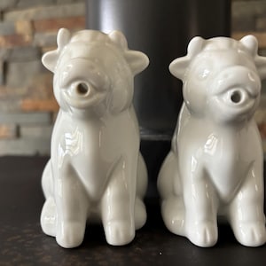 Może przedstawiać: Para białych ceramicznych solniczek w kształcie krów. Każda figurka ma błyszczące wykończenie, spiczaste uszy i mały okrągły otwór w okolicy pyska. Krowy przedstawiono w pozycji siedzącej, zwrócone przodem do widza, o gładkiej powierzchni.