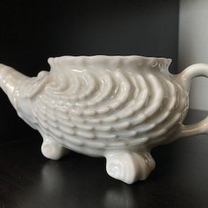 Puede incluir: Una tetera de cerámica blanca con forma de concha marina, con una superficie texturizada y tres patas. La tetera tiene un pico y una asa.