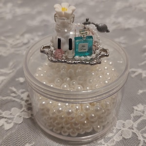 May include: A clear plastic container filled with white faux pearls. A small silver tray on top holds miniature perfume bottles, nail polish, and a flower-topped bottle. A string of pearls and a perfume atomizer complete the arrangement.
