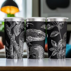 May include: Three stainless steel tumblers with clear lids, featuring a black and white line art illustration of a snake coiled among detailed botanical leaves and branches. The design wraps around the cylindrical bodies of the drinkware.