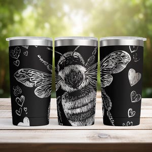 May include: Three stainless steel travel tumblers with a black background. Each tumbler features a detailed silver illustration of a bee. The tumblers also have silver hearts and a silver lid. The tumblers are on a wooden surface.
