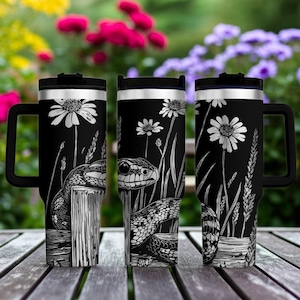 May include: A black stainless steel tumbler with a handle and a black lid, featuring a white line art illustration of a snake coiled around a wooden post amidst tall grass and daisy flowers, presented in three views to show the wrap-around design.