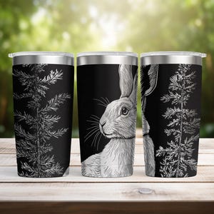 May include: Three stainless steel travel mugs with a black background. Each mug features a white illustration of a rabbit and a tree branch. The rabbit is depicted in profile, with detailed fur and whiskers. The tree branch is a detailed, white, botanical illustration.