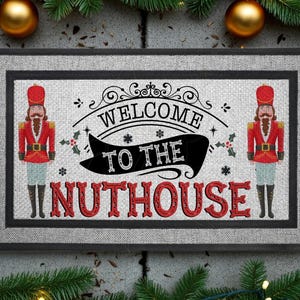 May include: A rectangular doormat with a light gray textured surface and a black border. It features two red and gold nutcracker figures on the sides and the black text WELCOME TO THE NUTHOUSE in a decorative font with red lettering for NUTHOUSE.