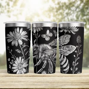 May include: Three black stainless steel tumblers with silver floral and bee designs. The tumblers feature detailed illustrations of daisies, butterflies, and a honeybee. The designs are etched onto the tumblers, creating a striking contrast.