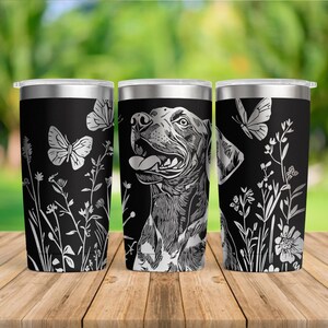 May include: Three black stainless steel tumblers with silver lids. Each tumbler features a different design: a dog portrait, butterflies, and floral elements. The tumblers are cylindrical and sit on a wooden surface.
