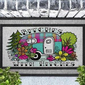May include: A rectangular doormat with a black border and a light gray center featuring a colorful illustration of a camper trailer, pine trees, and flowers. The text BITCHES WITH HITCHES is printed in black, bold, stylized lettering above and below.