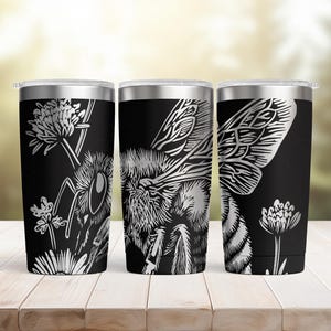 May include: Three stainless steel travel tumblers with a black background and a detailed white illustration of a bee and flowers. The tumblers have a silver rim and lid. The bee design is a detailed line drawing.