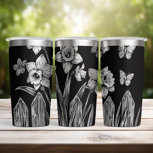May include: Three black insulated tumblers with silver lids. Each tumbler features a white floral design of daffodils, butterflies, and a picket fence. The tumblers are displayed on a wooden surface with a blurred green background.