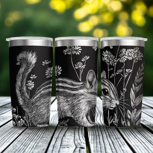 May include: A black stainless steel tumbler featuring a white line art illustration of a chipmunk surrounded by wildflowers and tall grasses. The design wraps around the cylindrical body, which is topped with a clear, removable plastic lid.