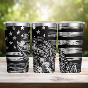 May include: Three stainless steel tumblers with a black and white design. The design features a frog illustration over a distressed American flag. The tumblers have a silver lid and are displayed on a wooden surface.