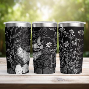 May include: Three stainless steel travel tumblers with black backgrounds. Each tumbler features a white illustration of a cat, flowers, and foliage. The cat's fur is detailed, and the flowers are delicate.