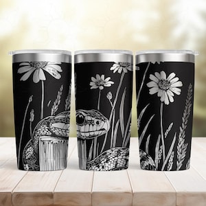 May include: Three black stainless steel tumblers with a white illustration of a snake, daisies, and tall grass. The snake is coiled around a wooden post. The tumblers have silver lids and are set on a wooden surface.