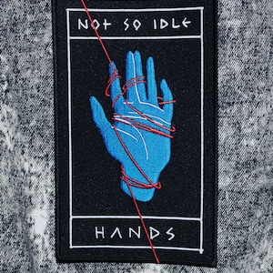 May include: A black and white embroidered patch with a blue hand tied with red string. The text "NOT SO IDLE HANDS" is written in white on a black background.