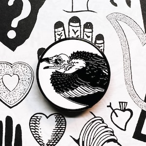 Peut inclure: Épinglette en émail noir et blanc représentant une illustration stylisée d'un vautour avec ses ailes déployées. L'épingle est circulaire et a un fond blanc.