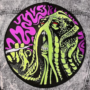 Op de afbeelding: Een zwarte en groene patch met een psychedelisch ontwerp van een wezen met een grote mond en een draaiend patroon. Het wezen wordt omringd door een paars en wit draaiend patroon.