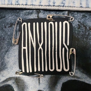 Könnte beinhalten: Ein schwarzer Stoff-Patch mit dem Wort "ANXIOUS" in weiß, befestigt an einer Jeansjacke mit Sicherheitsnadeln.