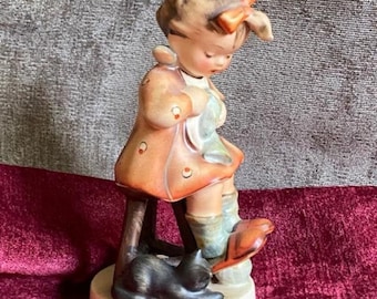 Puede incluir: Una figurita de porcelana de una niña con un vestido rojo y blanco sentada en un taburete con un gato negro a sus pies. La niña sostiene una muñeca y lleva un lazo rojo en el pelo.