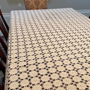 Pode incluir: Uma toalha de mesa de crochê branca com um delicado padrão floral. A toalha de mesa está colocada sobre uma mesa de madeira com uma cadeira no fundo.