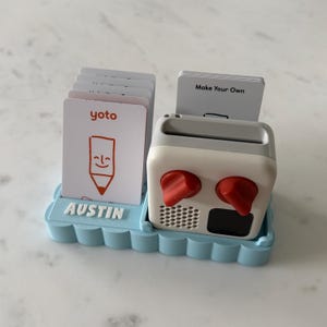 May include: A white and grey audio player with two red knobs, a speaker, and a small black screen. It sits in a light blue tray with the name "AUSTIN" and a stack of cards with the "yoto" logo and a smiling pencil.
