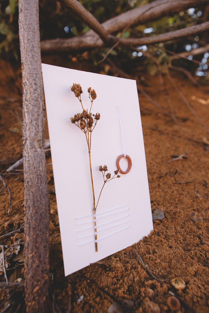 May include: A white card with a dried flower and a small terracotta ring attached with white thread. The card has white stitching across the bottom.