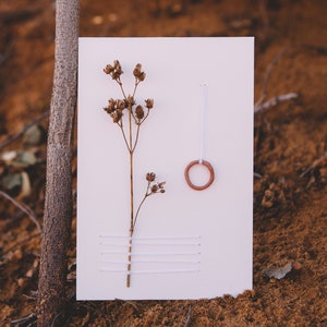 May include: A white card with a dried flower, a small terracotta ring, and white thread stitched in a horizontal pattern.