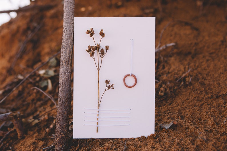 May include: A white card with a dried flower, a small terracotta ring, and white thread stitched in a horizontal pattern.