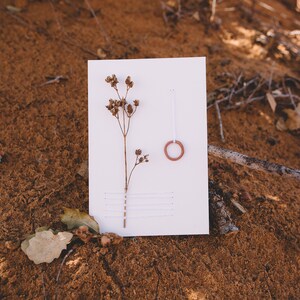 May include: A white card with a dried flower and a small terracotta ring on a brown sandy surface.