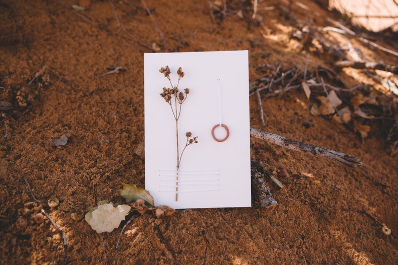 May include: A white card with a dried flower and a small terracotta ring on a brown sandy surface.