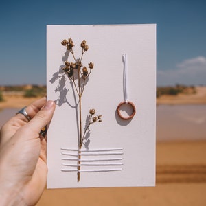 May include: A white card with a dried flower, white thread, and a small copper ring. The card is held in a hand against a blurred background of sand and water.