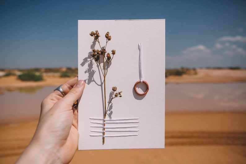 May include: A white card with a dried flower, white thread, and a small copper ring. The card is held in a hand against a blurred background of sand and water.