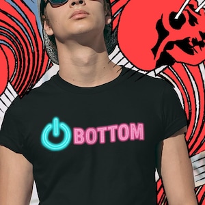 Könnte beinhalten: Schwarzes T-Shirt mit dem Wort "BOTTOM" in Pink und einem Power-Button-Symbol in Türkis. Der Hintergrund zeigt ein rot-weißes Design mit roten Totenköpfen und Blitzen.