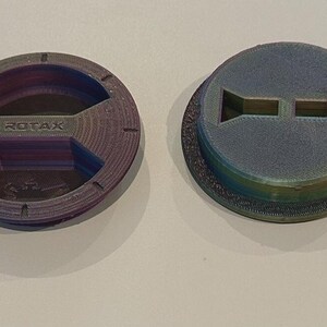 Puede incluir: Dos piezas circulares de pl&aacute;stico multicolor. Una pieza tiene la palabra "ROTAX" grabada en el centro. La otra pieza tiene dos recortes rectangulares. Ambas piezas tienen un acabado texturizado e iridiscente, con tonos morados, azules y verdes.