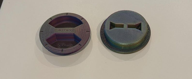 Puede incluir: Dos piezas circulares de pl&aacute;stico multicolor. Una pieza tiene la palabra "ROTAX" grabada en el centro. La otra pieza tiene dos recortes rectangulares. Ambas piezas tienen un acabado texturizado e iridiscente, con tonos morados, azules y verdes.