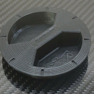 Puede incluir: Un objeto negro circular impreso en 3D con un dise&ntilde;o segmentado. Presenta un recorte central en forma de cruz y una secci&oacute;n curva. Peque&ntilde;as hendiduras est&aacute;n espaciadas alrededor del borde exterior. La superficie tiene una apariencia texturizada.