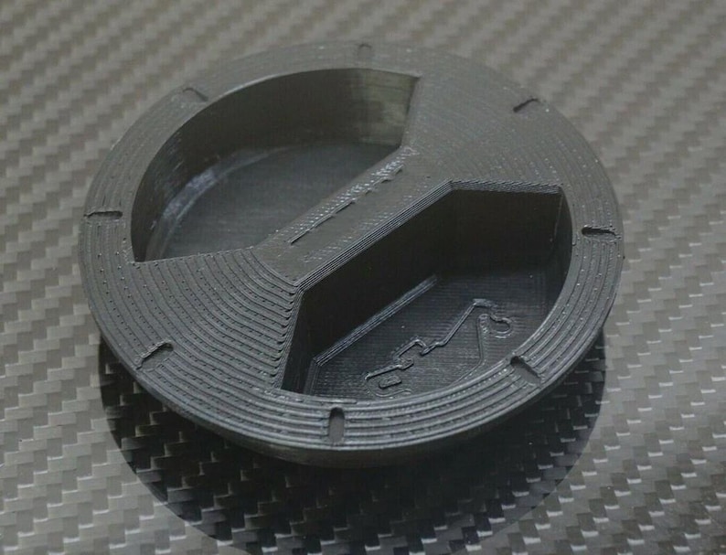 Puede incluir: Un objeto negro circular impreso en 3D con un dise&ntilde;o segmentado. Presenta un recorte central en forma de cruz y una secci&oacute;n curva. Peque&ntilde;as hendiduras est&aacute;n espaciadas alrededor del borde exterior. La superficie tiene una apariencia texturizada.