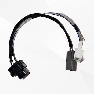Puede incluir: Un arnés de cableado automotriz que presenta un conector de plástico negro en un extremo y dos conectores separados, uno gris y otro blanco, en el otro extremo. Los cables están agrupados con una funda protectora de malla negra.