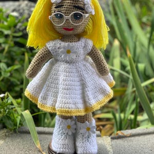 May include: A crocheted doll with yellow hair, wearing a white dress with yellow trim and white shoes with daisy accents. The doll is wearing a white headband with daisy accents and round glasses.