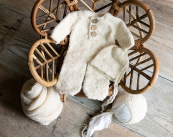 May include: A white knitted baby outfit with a matching hat and a wooden chair. The outfit has a button closure and the hat has a simple design.