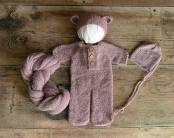 May include: A mauve knitted baby outfit with a bear hat, a matching hat, and a knitted wrap. The outfit has wooden buttons.