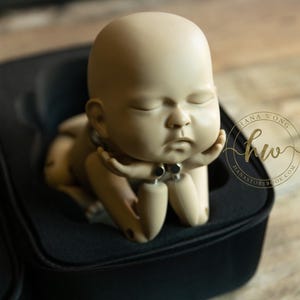 May include: A beige-colored baby doll with closed eyes, resting its head on its hand. The doll is inside a black case. The doll has a small silver necklace. The image includes the text "Hana Wong" and "hanastorybook.com".