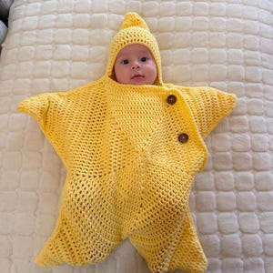 May include: A bright yellow crocheted star-shaped bunting with a matching pointed hood. The garment features a wide opening for the face and is secured at the front with two dark brown circular buttons, creating a cosy, five-pointed star silhouette.