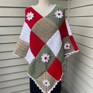 May include: A handmade crochet poncho in a patchwork design. The poncho features squares of red, white, olive green, and tan. White daisy-shaped appliques are sewn on the front. The edges are trimmed with white scalloped edging.