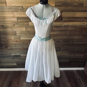 May include: A white dress with a light blue sash and accents, displayed on a mannequin. The bodice features a textured pattern and a ruffled neckline with a small bow. The skirt is full and flowing, creating a vintage or costume aesthetic.