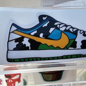 May include: A white, black, and blue sneaker with a cow print design and a yellow swoosh. The sneaker has a green and blue trim around the sole.