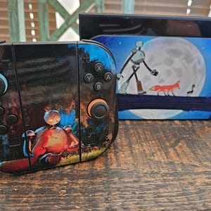 May include: A black and blue Nintendo Switch controller and dock set. The controller features a robot illustration with orange and red accents. The dock has a full moon and fox scene. The set is on a wooden surface.