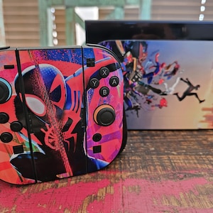 May include: A colorful Nintendo Switch Joy-Con controller and console featuring a Spider-Man graphic. The controller is predominantly pink and red with black buttons. The console has a matching design with a cityscape background.