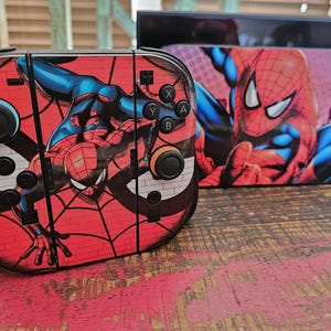 May include: A red and black Spider-Man themed Nintendo Switch console and dock. The console controllers feature the superhero on a brick wall background. The dock has a large image of Spider-Man. The set is on a wooden surface.