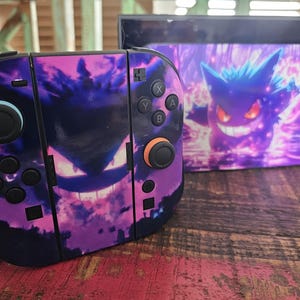 May include: A Nintendo Switch console and dock set with a purple and black Gengar design. The Joy-Cons feature a Gengar face, while the dock displays a Gengar illustration. The set is on a wooden surface.